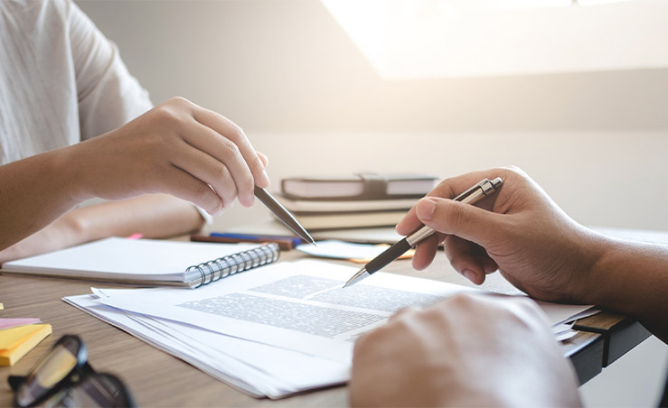 Two people pointing pens above paper on desk