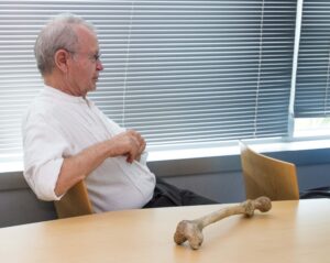 Thomas Quinn, D.O. sitting in chair with human bone on table looking towards front of room